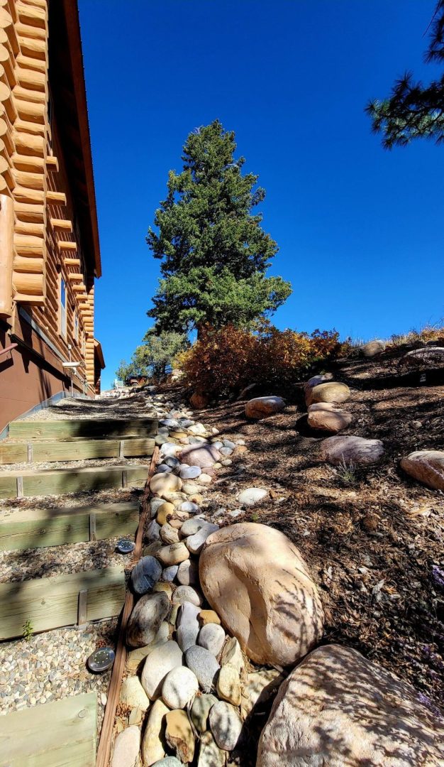 A log cabin on a hillside with rocks and a tree.